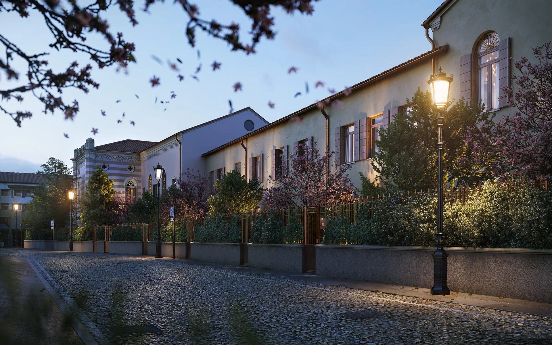 Strada acciottolata serale con lampioni illuminati e edificio storico con finestre ad archi e giardino fiorito.