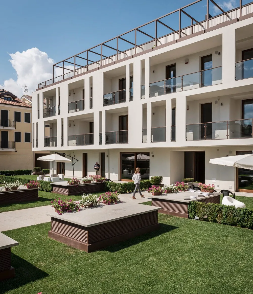 Giardino urbano con siepi curate, fioriere e poltrone in uno spazio all'aperto davanti a un edificio moderno a più piani con balconi in vetro.