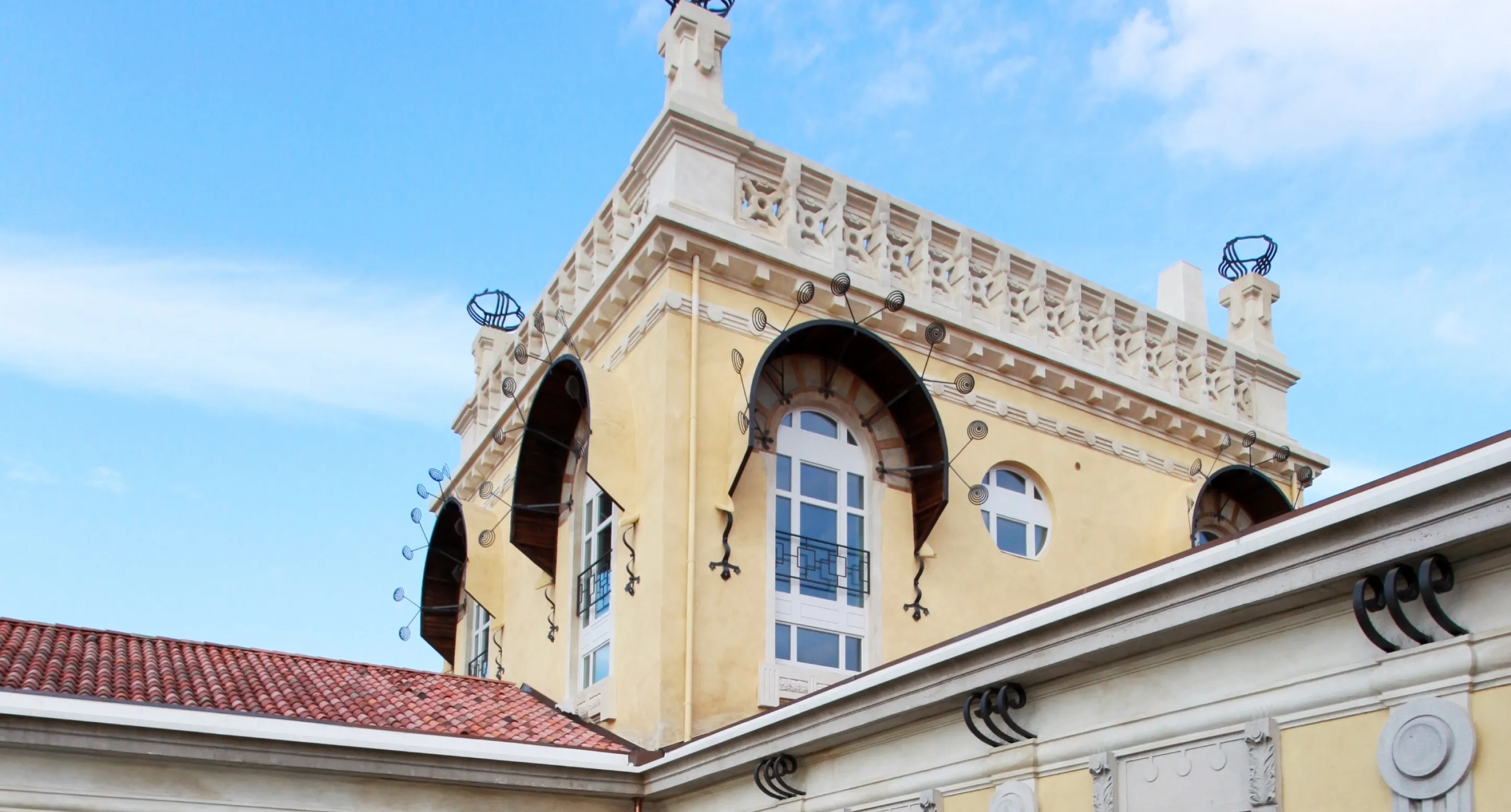 Dettaglio di un edificio storico con finestre ad arco, verande ornamentali in ferro battuto e cornici decorative sotto un cielo azzurro.