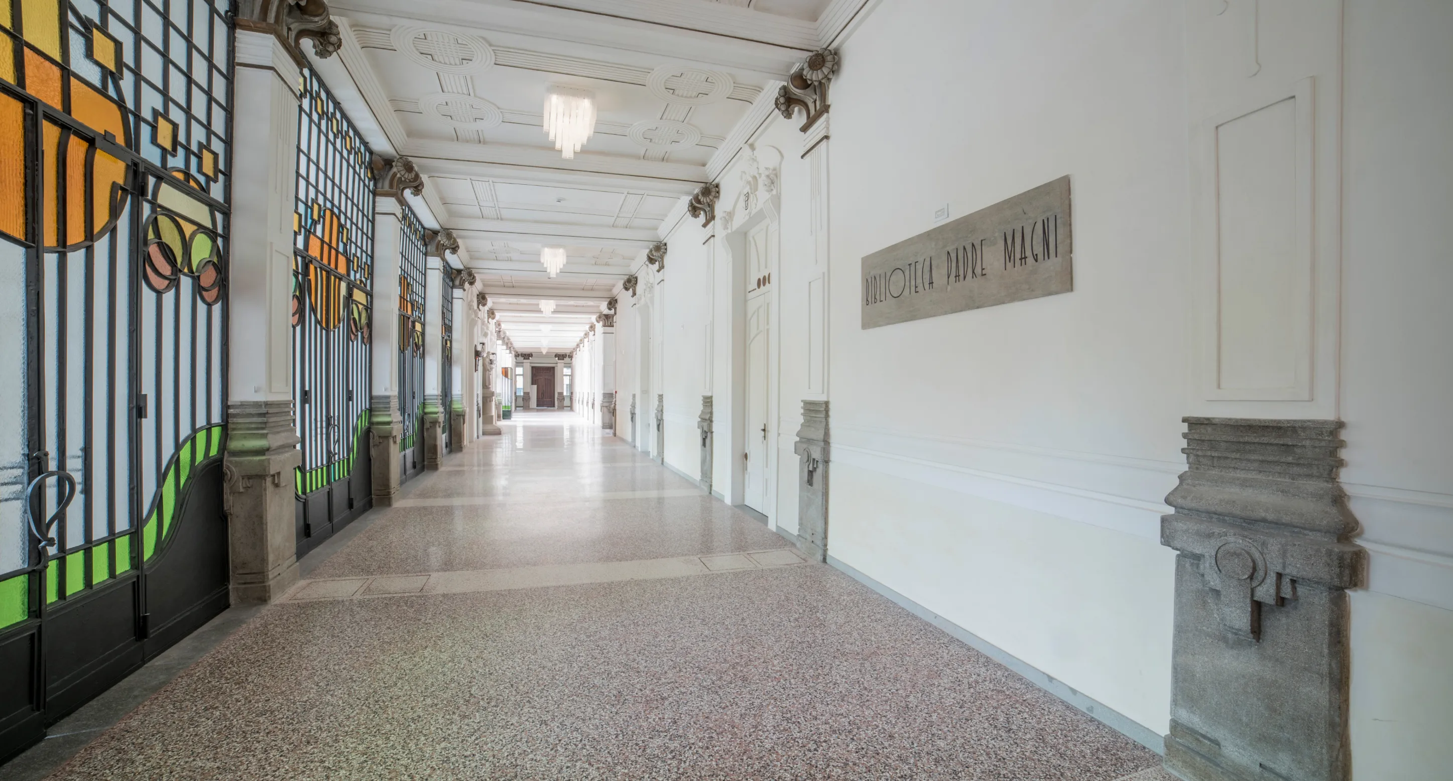 Corridoio luminoso con porte a vetri colorate sulla sinistra e una targa a destra con scritto Biblioteca Padre Macni.