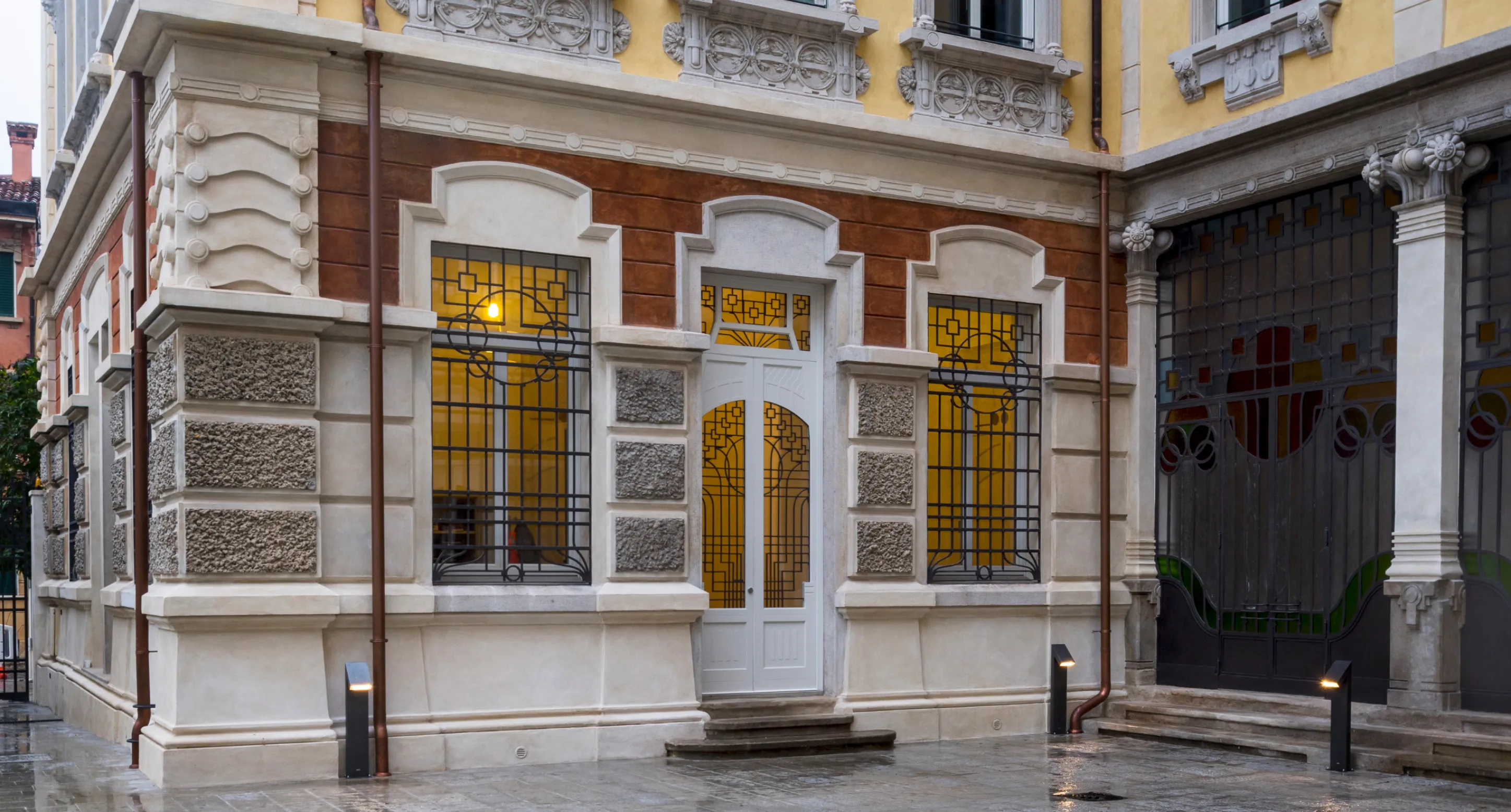 Ingresso con porta bianca e finestre decorate con griglie di ferro in un edificio storico con facciata in pietra e mattoni.