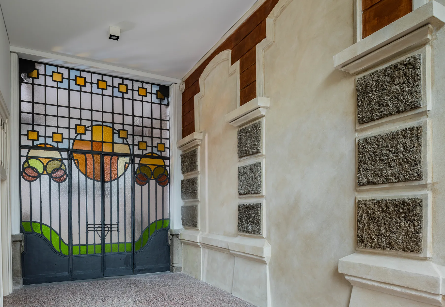 Ingresso con porta in ferro battuto e vetro colorato con motivo geometrico e cerchi arancioni, pareti beige con decorazioni in rilievo e finiture in pietra.