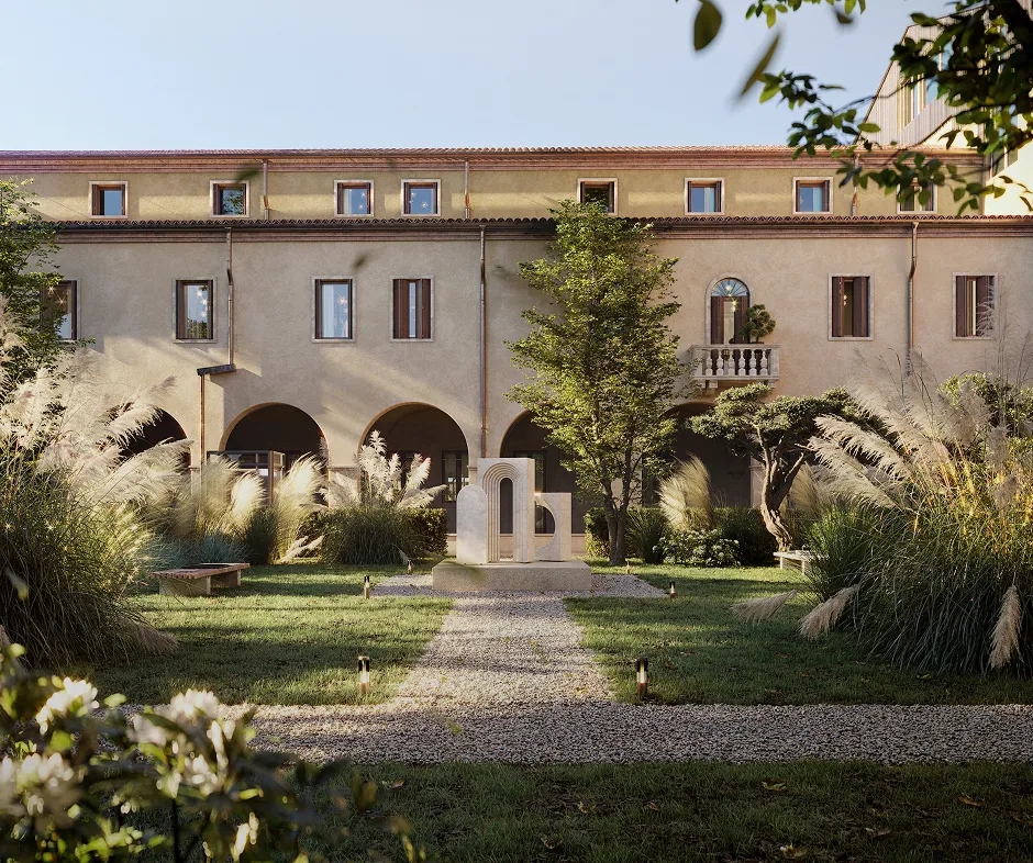 Stone courtyard with gravel pathways, ornamental grasses, trees, and a modern abstract sculpture in front of a beige building with arched doorways and multiple windows.
