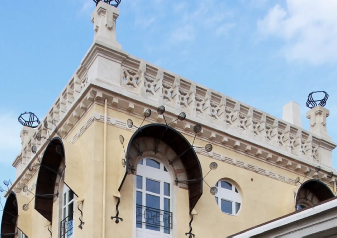 Dettaglio della parte superiore di un edificio storico con finestre ad arco, decorazioni in ferro battuto, e cielo blu nuvoloso sullo sfondo.
