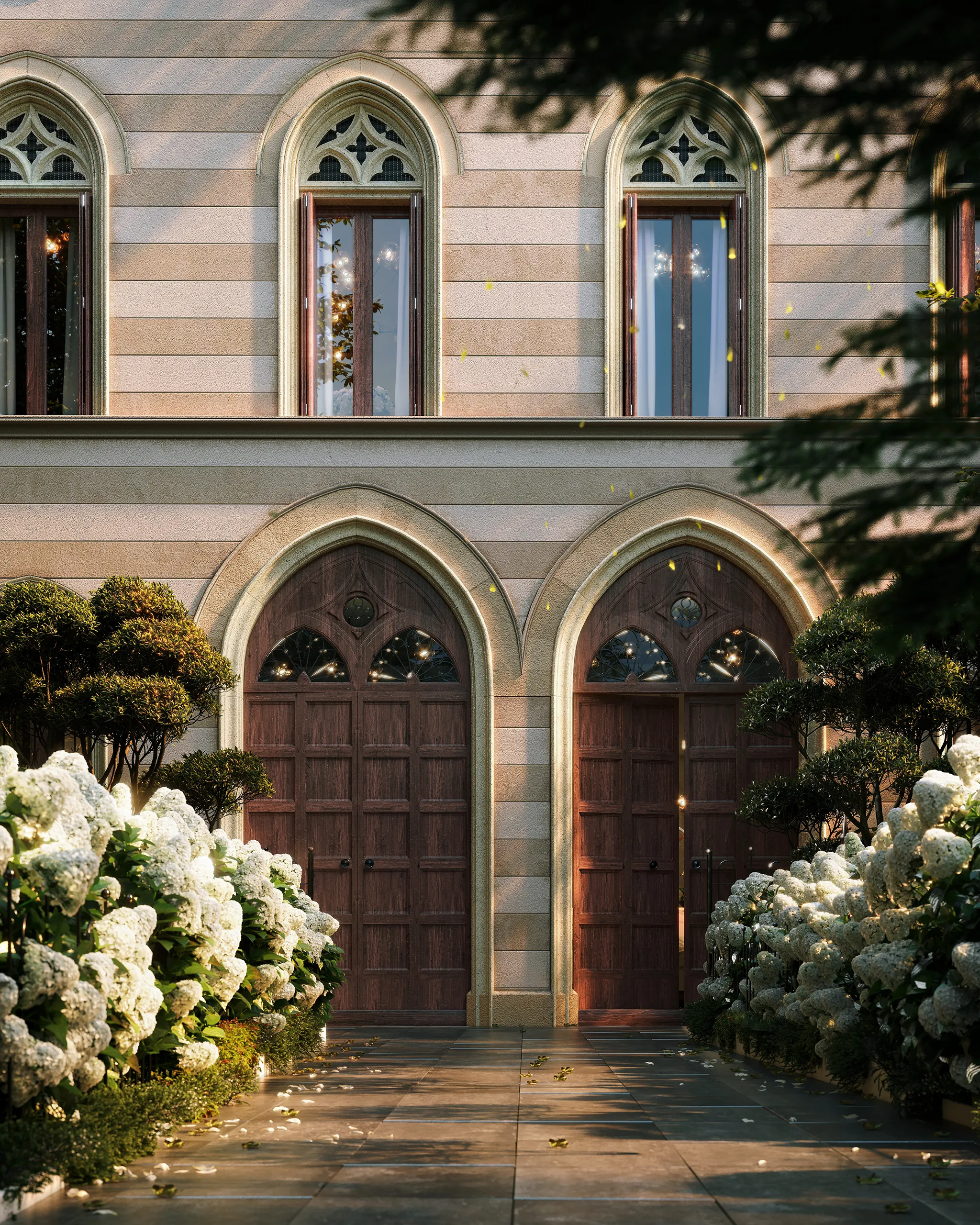 Pathway lined with white hydrangea flowers and trimmed bushes leading to two large wooden arched doors on a stone building with arched windows above.