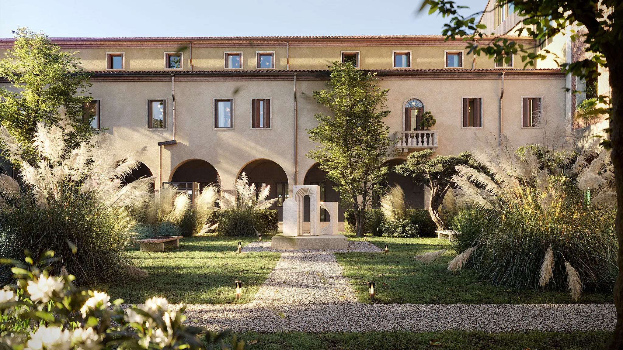 Giardino verde con alberi, cespugli e un percorso di ghiaia che conduce a una scultura astratta davanti a un edificio con archi e finestre.