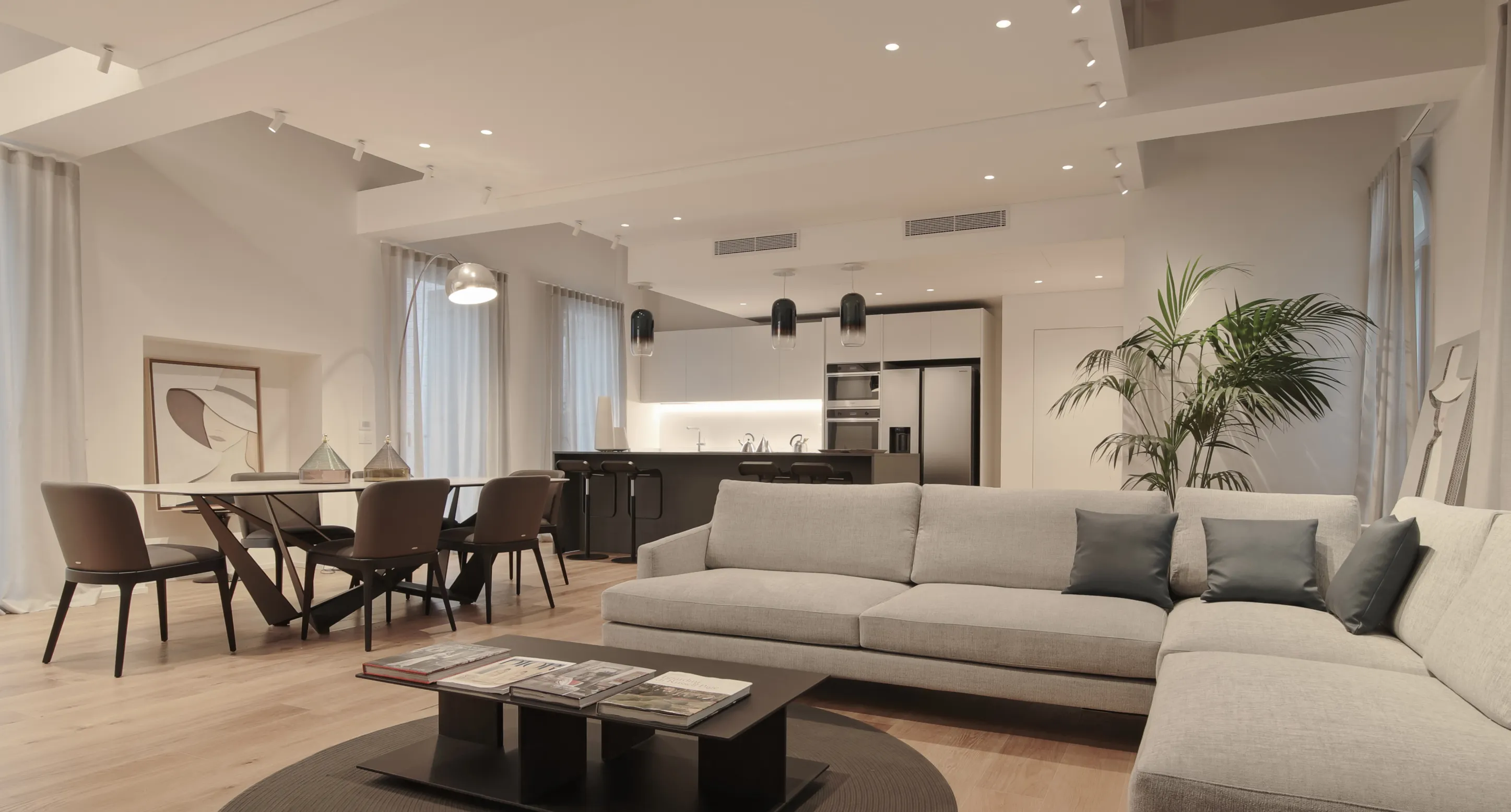 Soggiorno moderno con divano grigio angolare, tavolino basso con riviste, tavolo da pranzo con sedie scure e cucina aperta sullo sfondo.