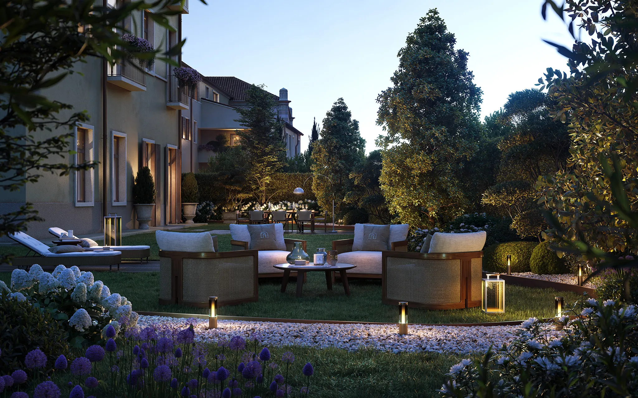 Spazio esterno di sera con mobili da giardino, lampade illuminate, fiori viola e bianchi e siepi curate.