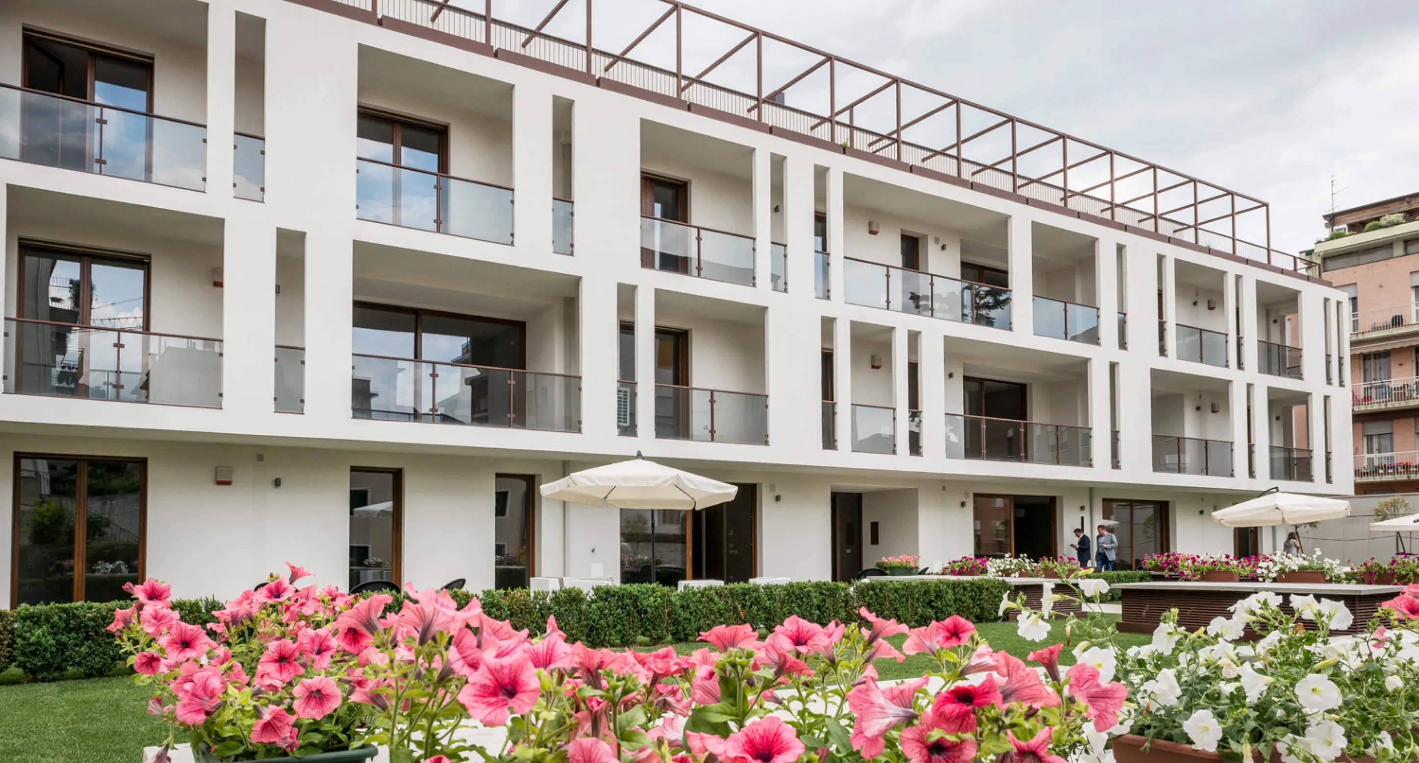 Edificio moderno con balconi in vetro e fiori rosa e bianchi in primo piano su un prato verde.