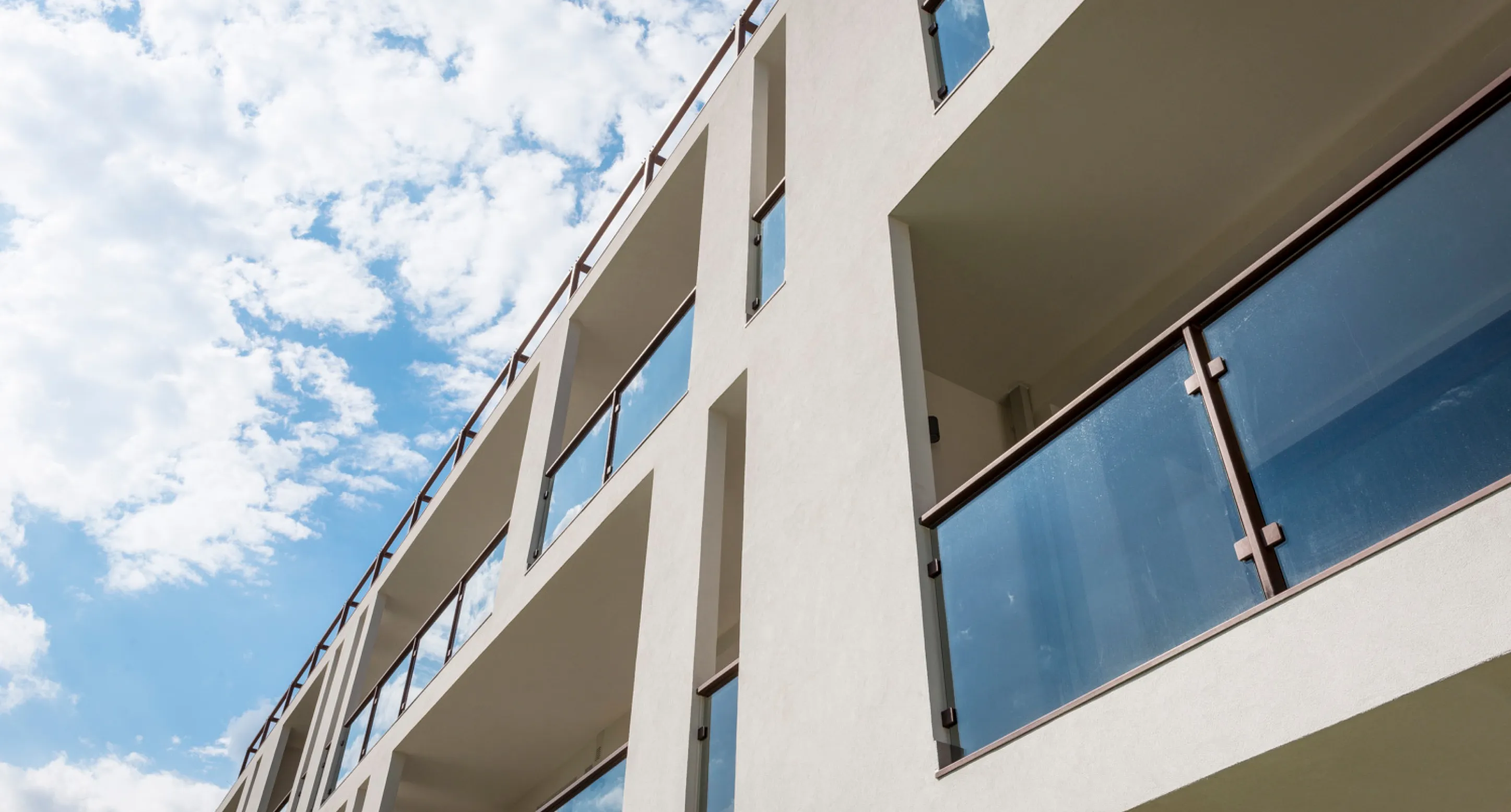 Vista verso l'alto di un edificio moderno con balconi dotati di parapetti in vetro sotto un cielo parzialmente nuvoloso.
