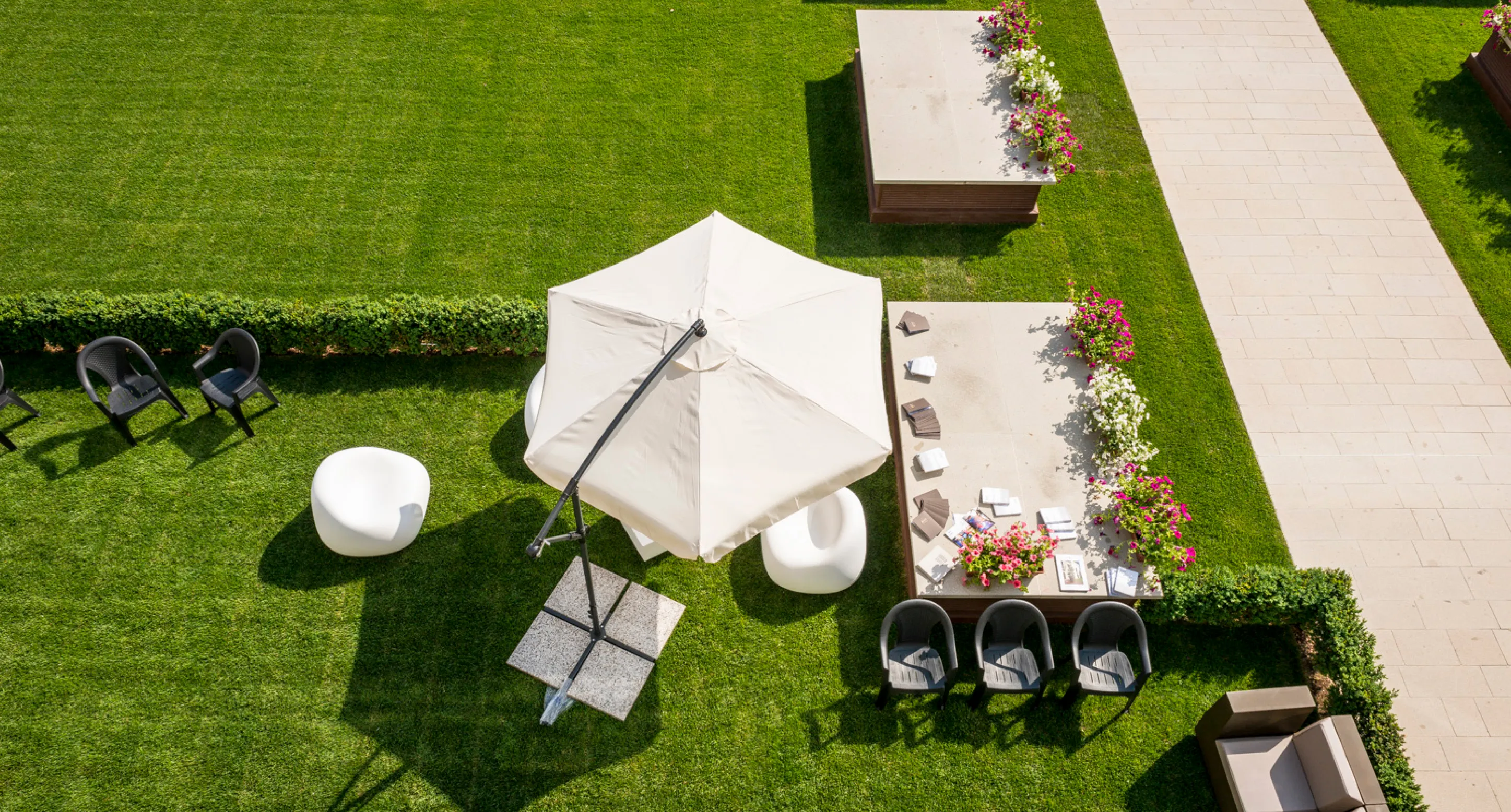 Area esterna con ombrellone beige, sedie nere e bianche, tavolo con libri e fiori su prato verde.