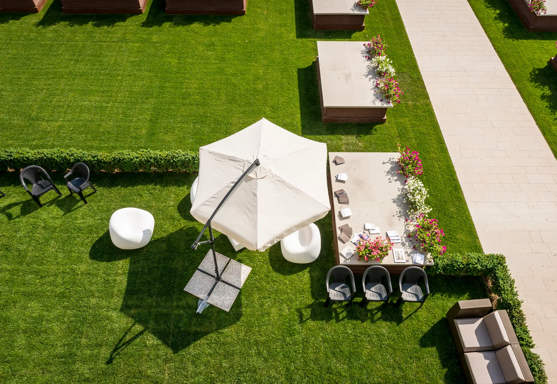 Vista dall'alto di un giardino con un grande ombrellone bianco, sedie moderne nere e bianche, tavoli decorati con fiori colorati e un camminamento in pietra.